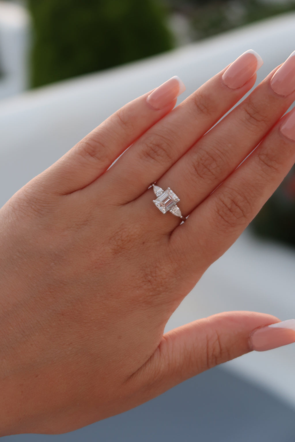 Emerald cut moissanite three-stone engagement ring with triangular side stones worn on hand with French manicure showcasing step cut center stone and elegant design