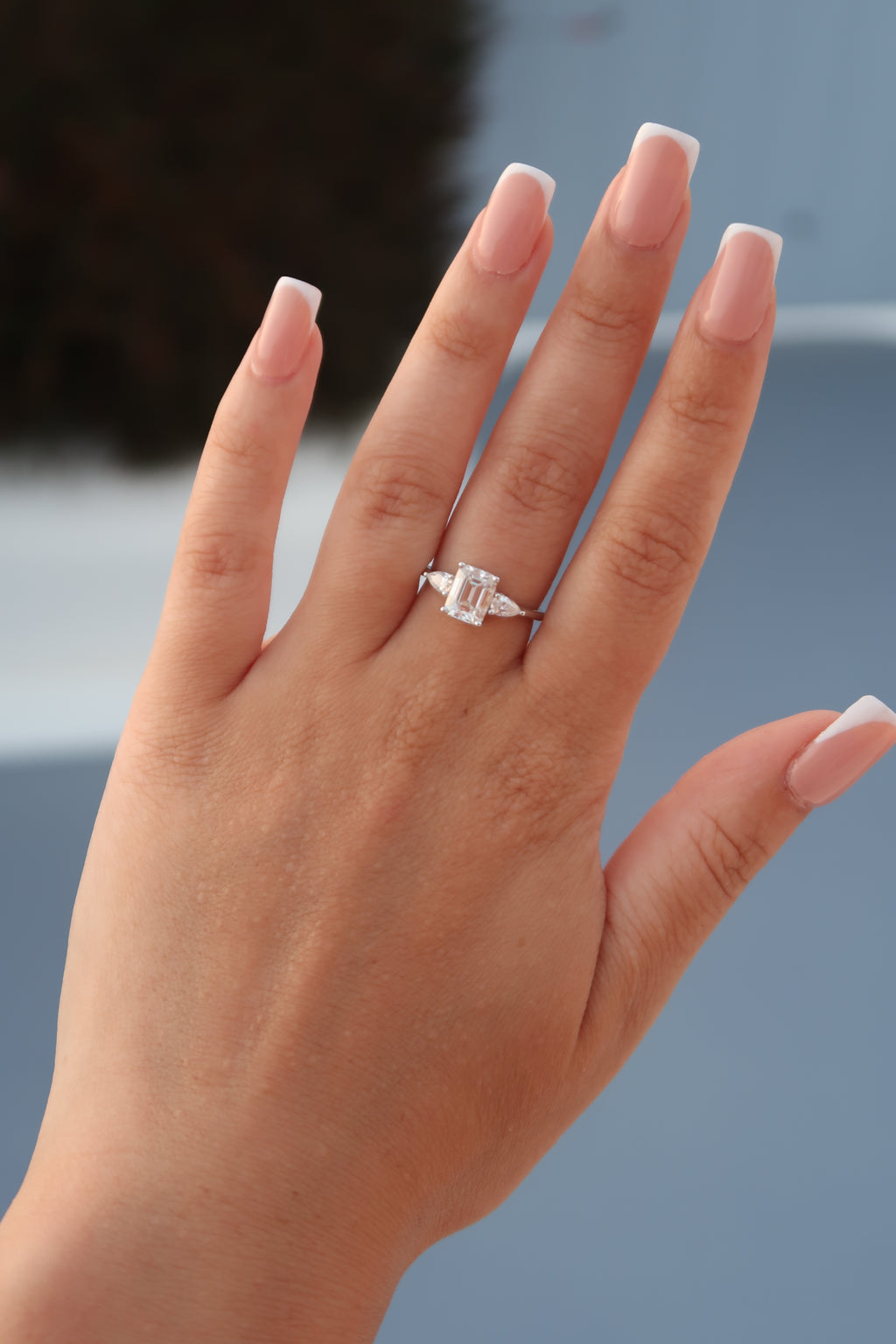Emerald cut moissanite three-stone engagement ring with triangular side stones worn on hand with French manicure showcasing step cut center stone and elegant design