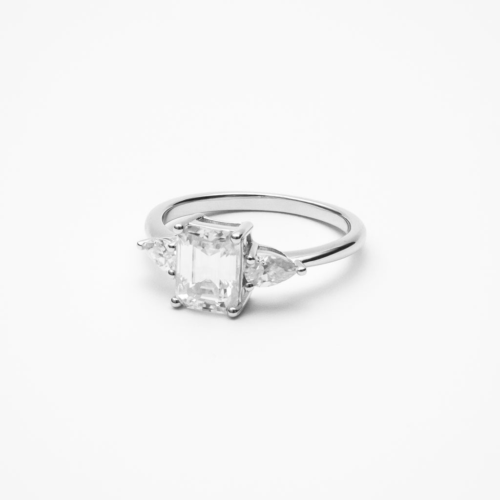 Emerald cut moissanite three-stone engagement ring with pear-shaped side stones in white gold setting on clean white background showcasing step cut center stone and elegant silhouette