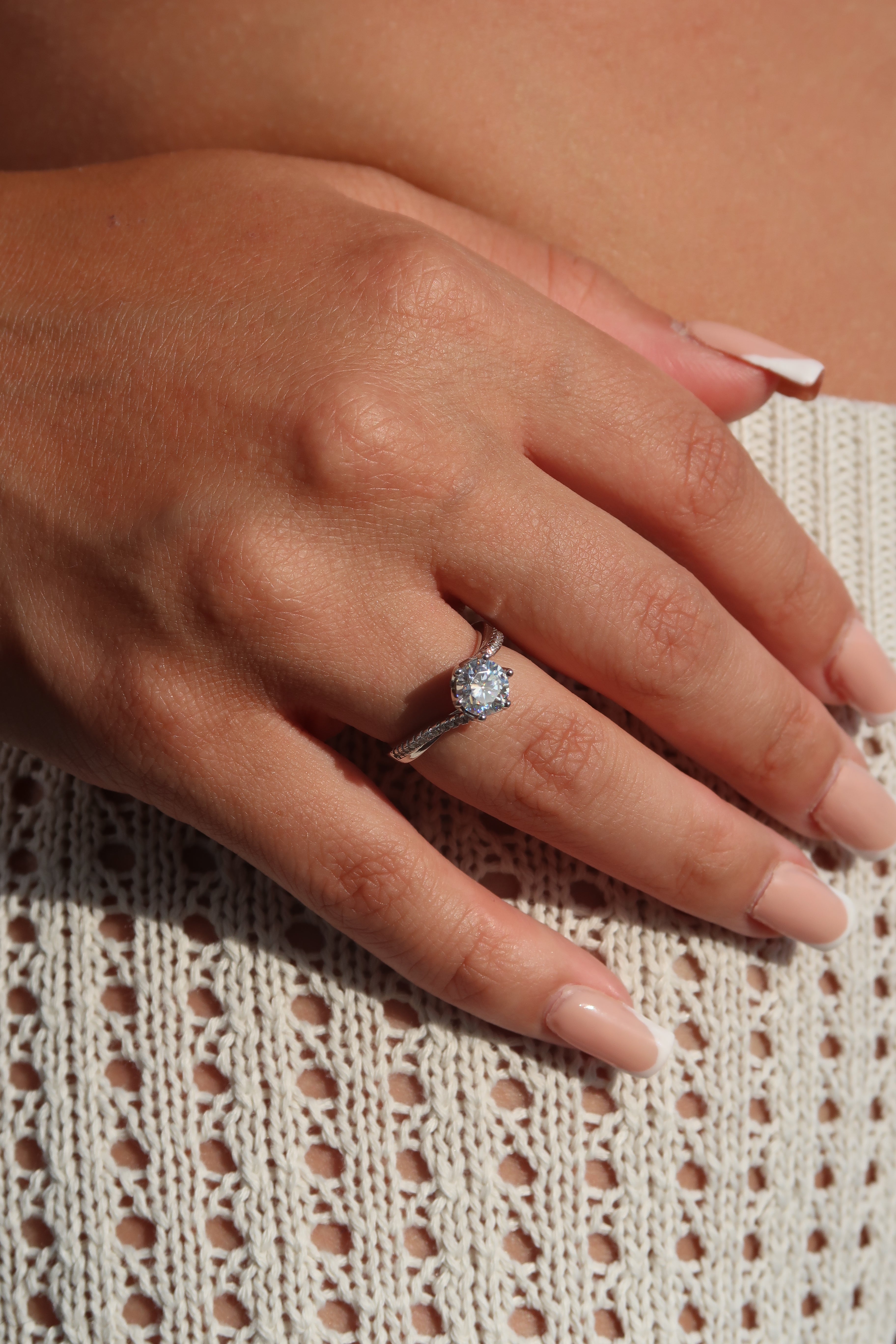 Hand wearing round cut moissanite engagement ring with twisted band on textured fabric highlighting elegant sparkle