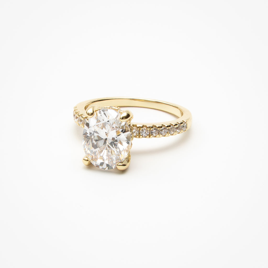 Oval cut cubic zirconia engagement ring with pavé band and hidden halo in yellow gold setting on clean white background