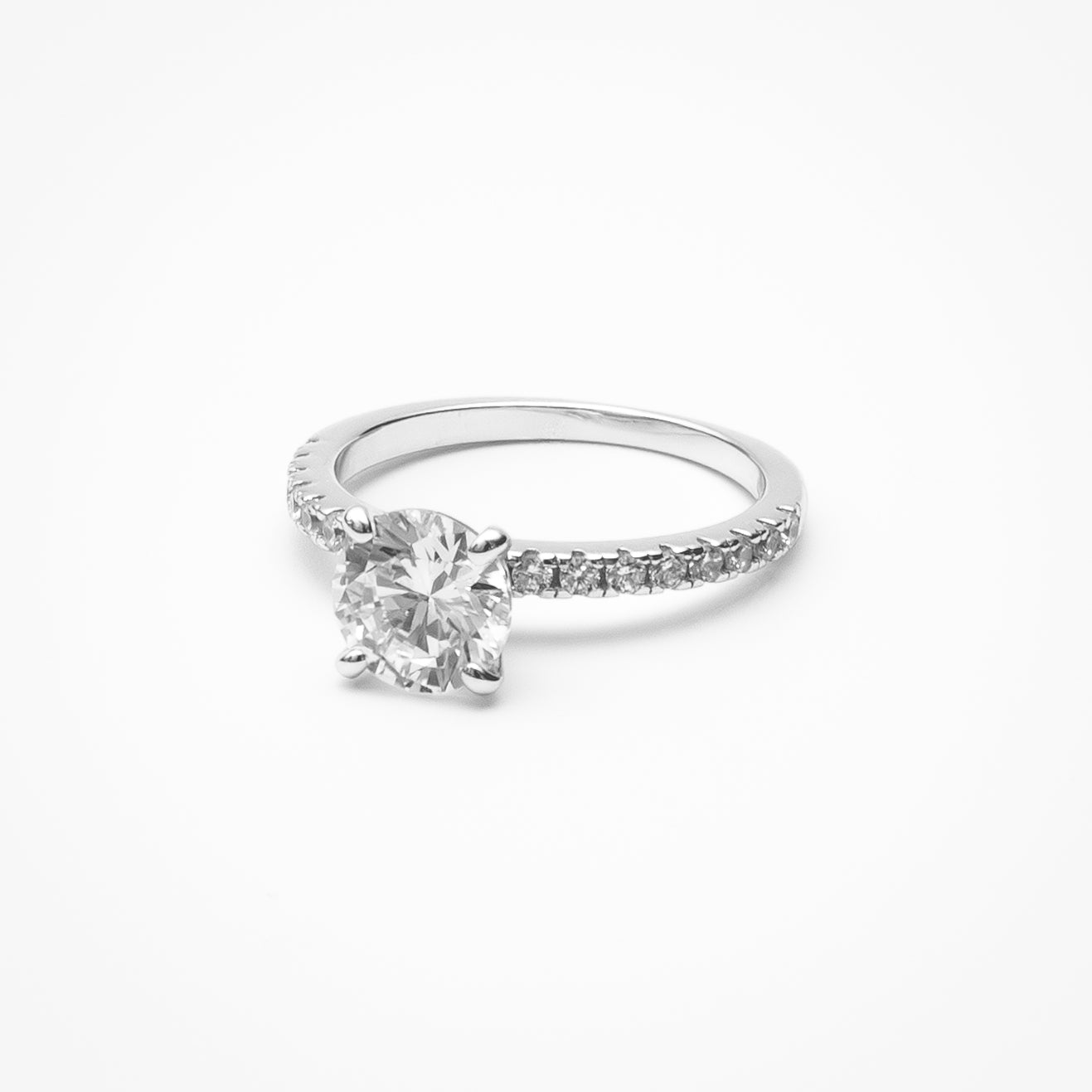 Round cut cubic zirconia engagement ring with pavé band in white gold setting on clean white background showcasing four-prong center setting