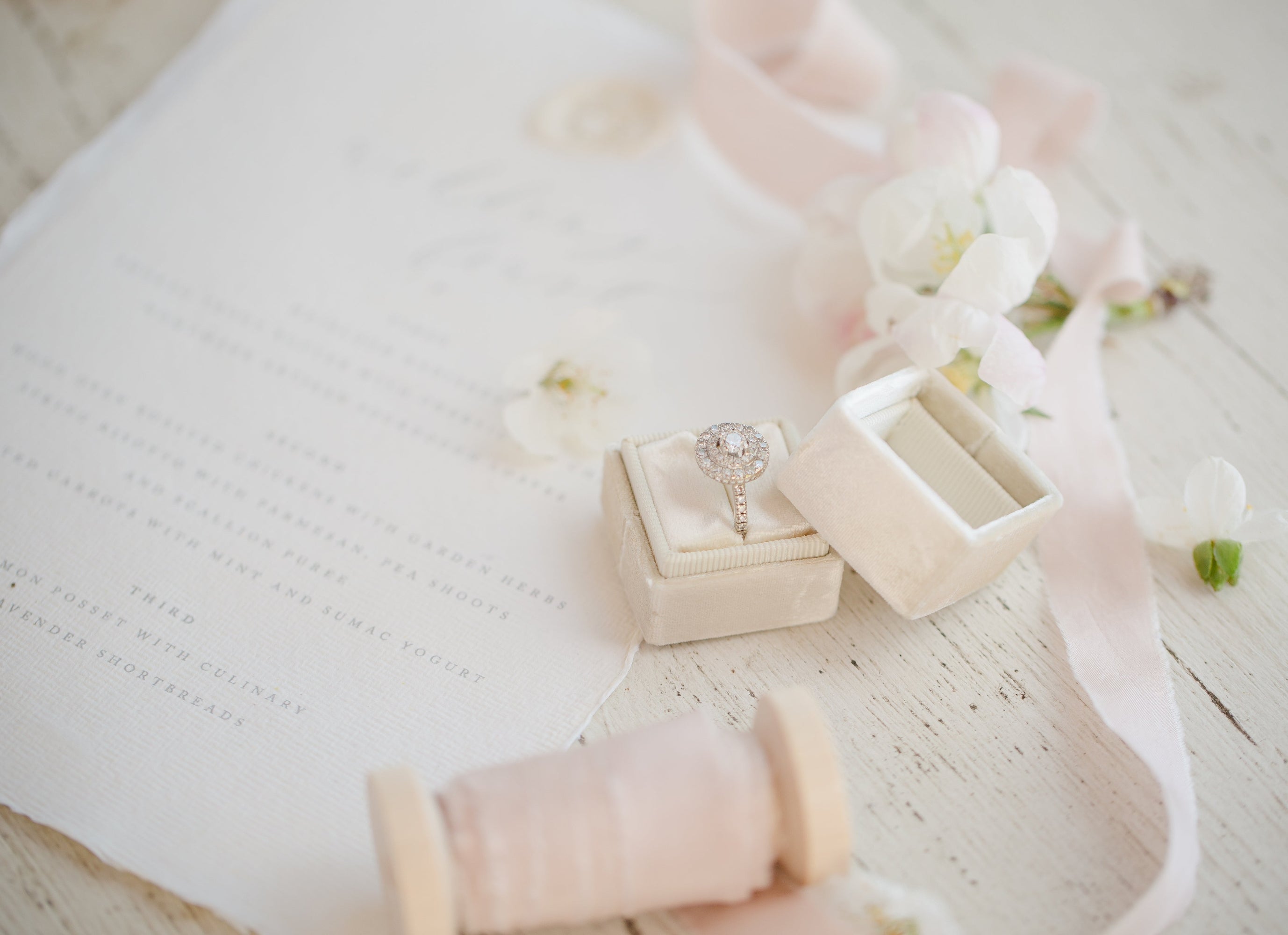 Round cut moissanite halo engagement ring displayed in cream velvet ring box surrounded by soft white flowers and romantic bridal styling on vintage white wood background