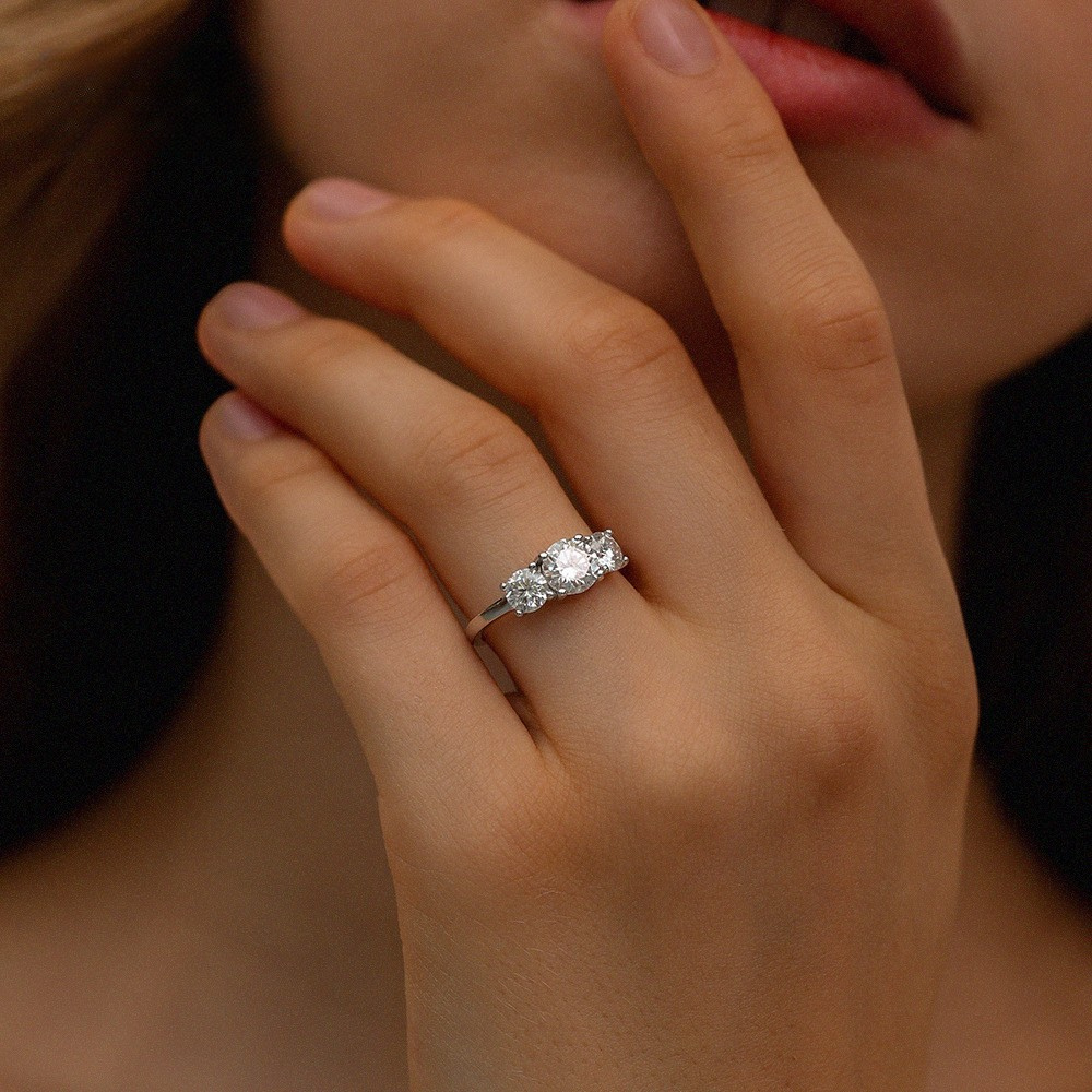 Hand wearing round cut moissanite three-stone engagement ring in dramatic lighting highlighting brilliant sparkle
