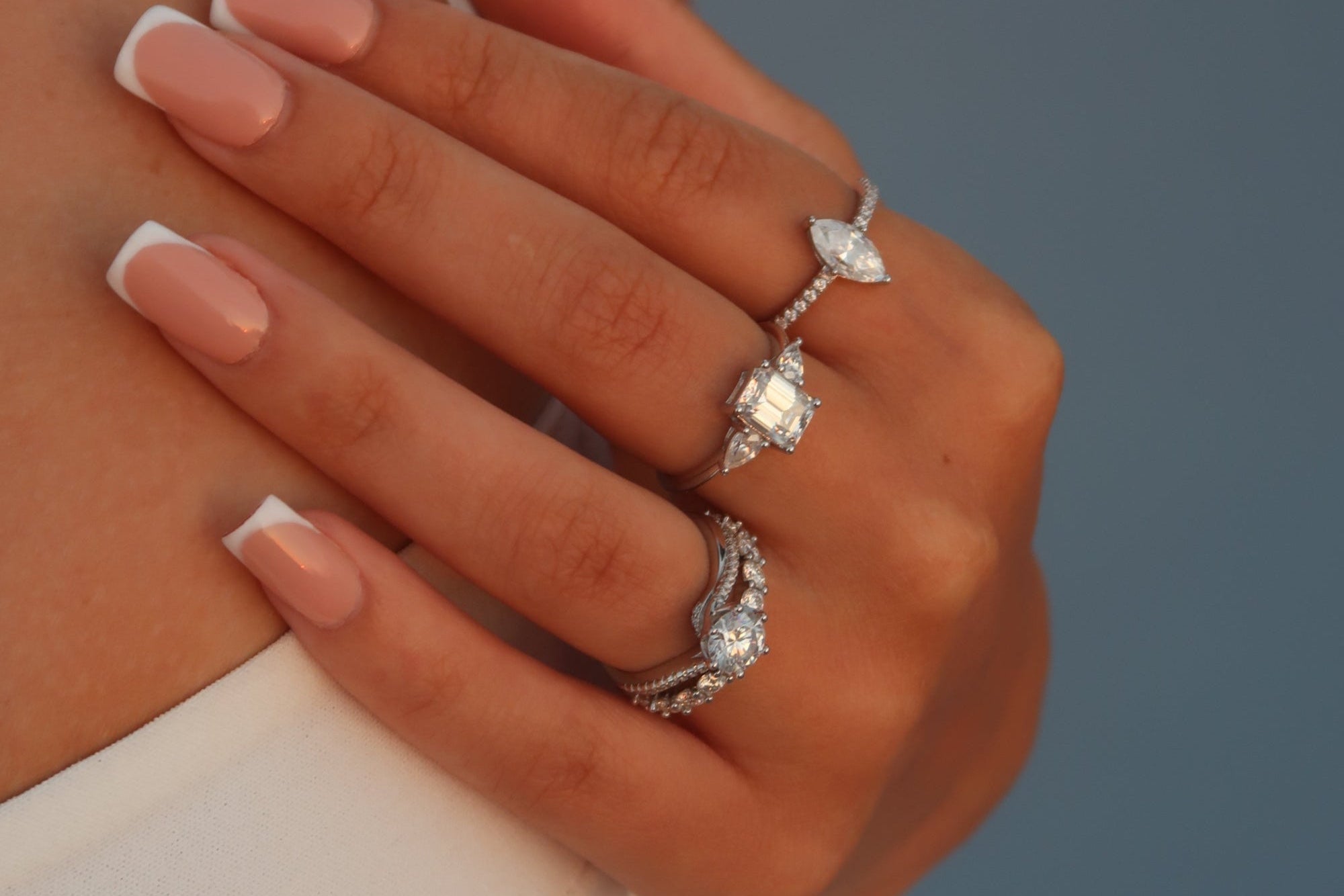 Three moissanite rings displayed on fingers with French manicure nails - featuring a marquise cut solitaire, an emerald cut center stone with side stones, and an eternity band with multiple round brilliant stones, all set in white metal