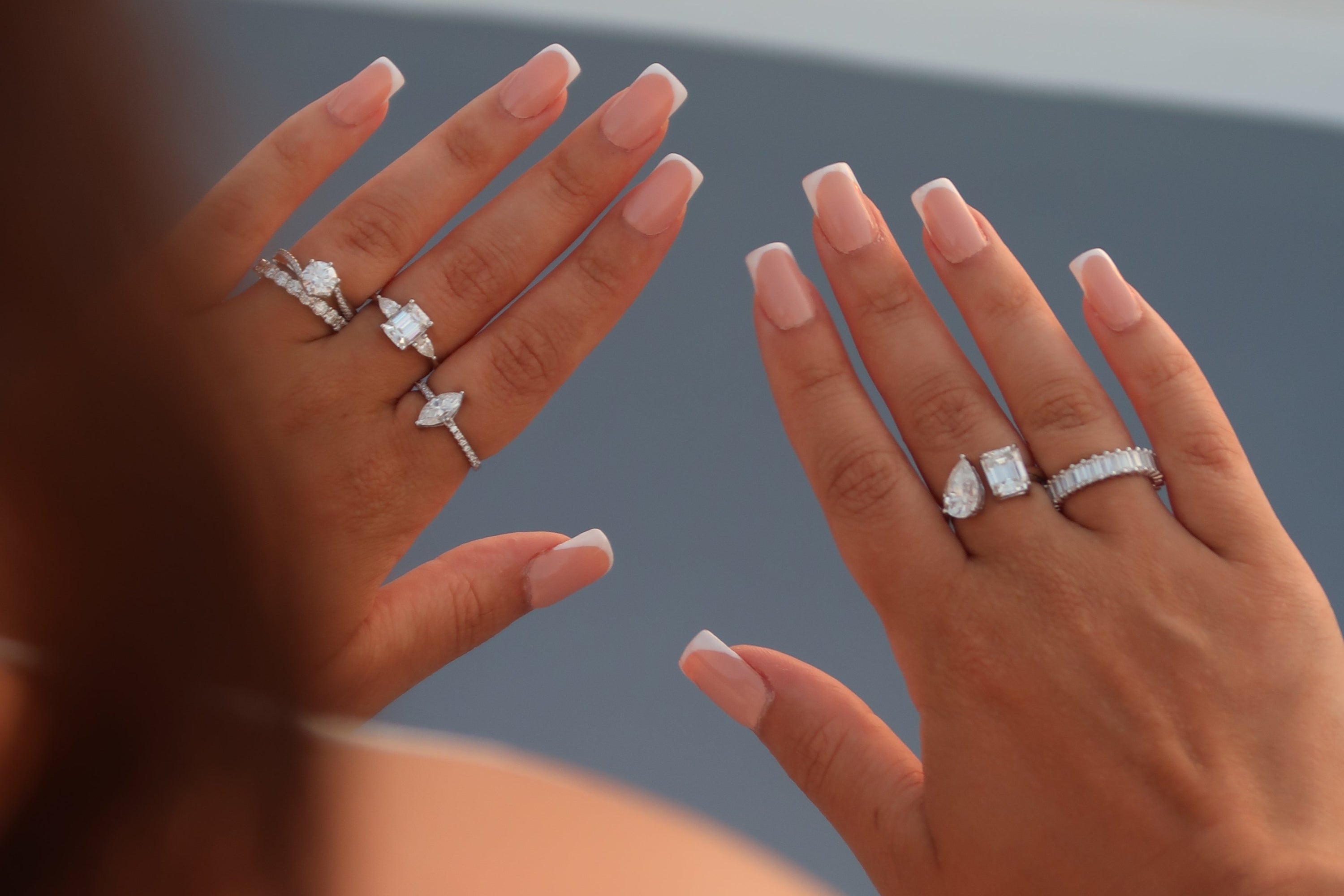 Two hands displaying collection of moissanite rings including emerald cut solitaires, marquise cut rings, oval and round brilliant stones in white gold settings with French manicure nails