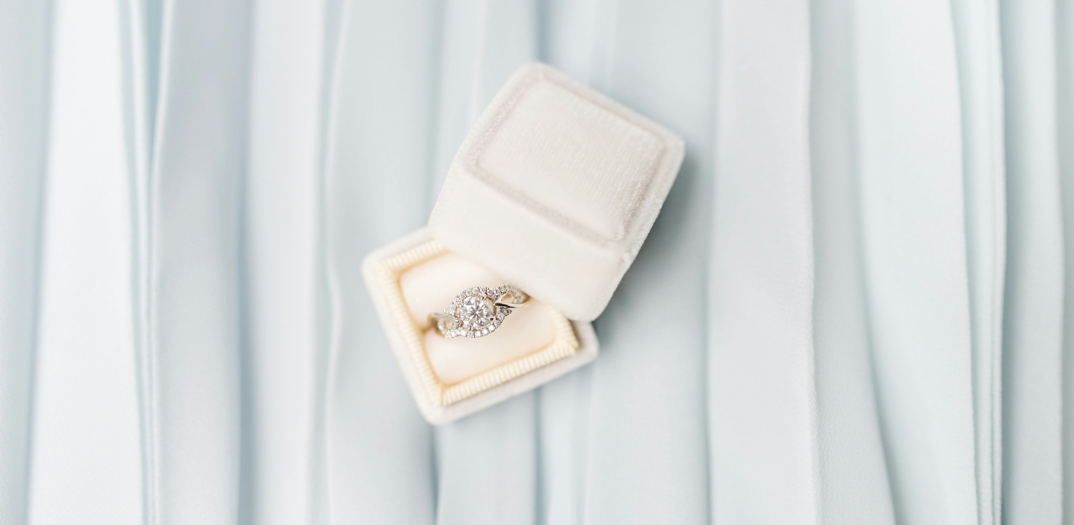 Moissanite engagement ring with round center stone and halo setting displayed in cream velvet ring box on white pleated fabric background