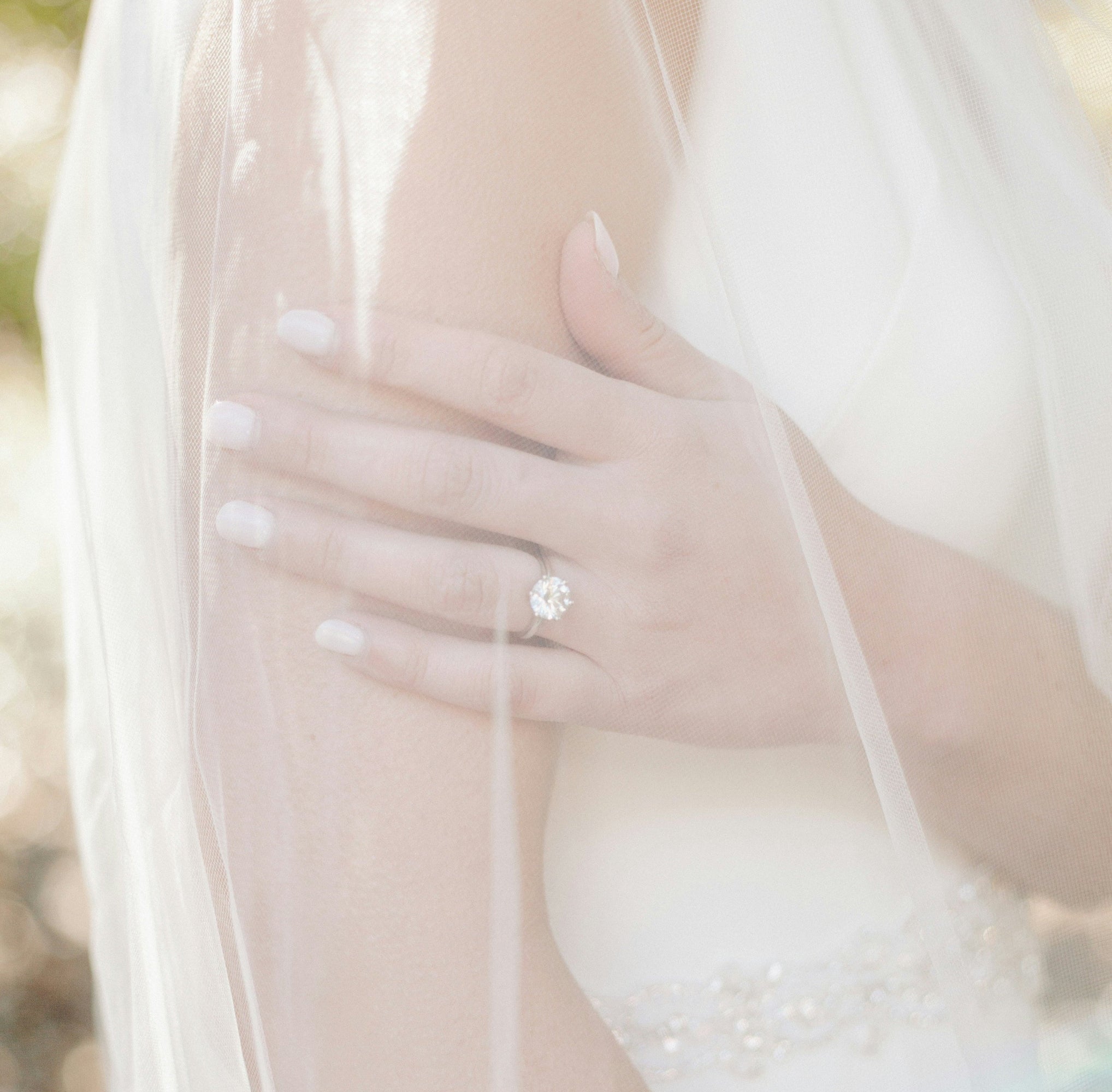 Moissanite solitaire engagement ring with round brilliant center stone worn with flowing white bridal veil in soft romantic lighting